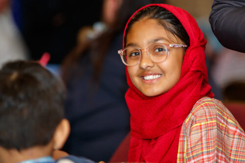 A girl with glasses and a red headscarf smiling at the camera