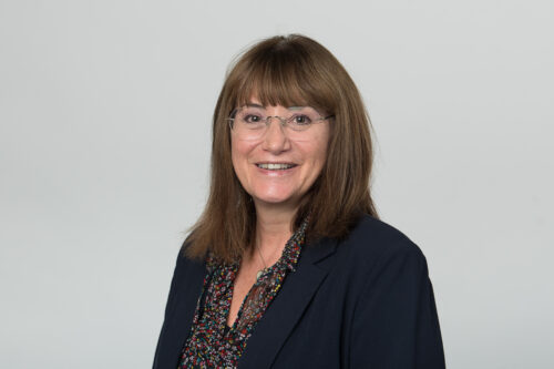 Chief executive Jo Savage wearing a black shirt and black jacket, with the grey background smiling at the camera