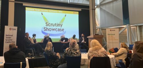 Six people sitting on chairs on a stage in front of an audience.  The screen behind reads 'scrutiny showcase'.